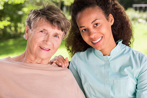 Young nurse in teal scrubs smiling next to elderly woman outside